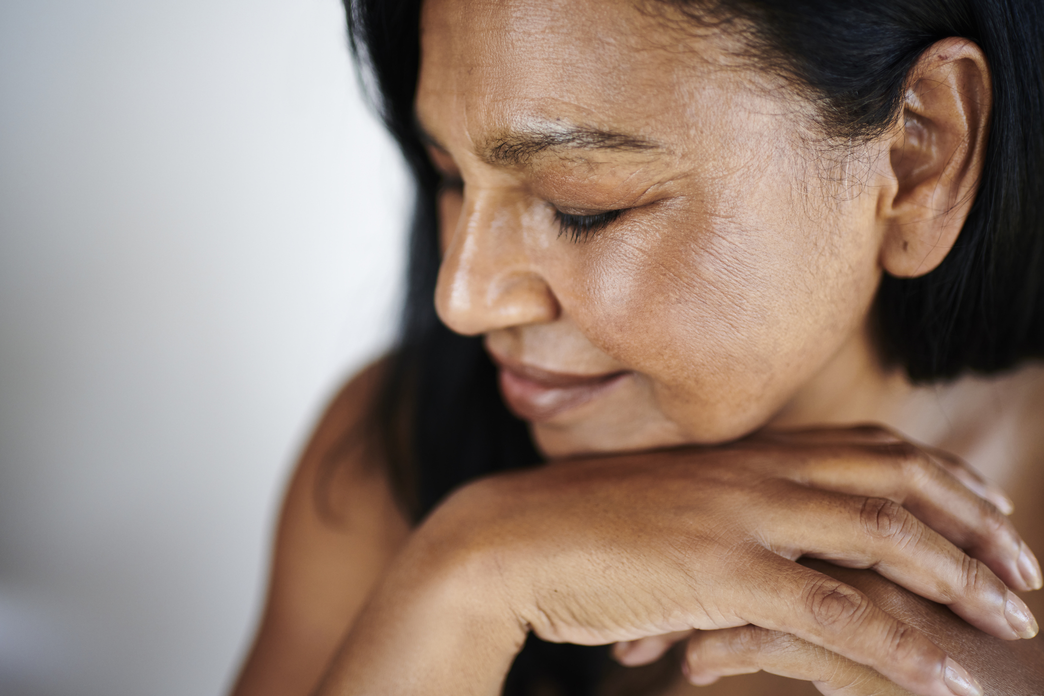 A serene mature woman of South Asian ethnicity sits with her eyes closed, hands gently resting on her chin, radiating calm and contentment in soft natural light.