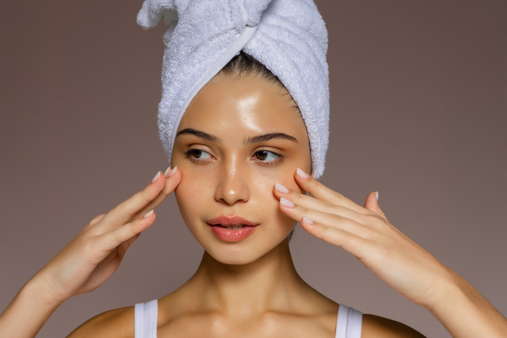 Young woman massaging her face with both hands after a shower, wearing a towel on her head and showing a fresh skincare routine